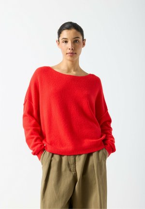 Donna con capelli scuri legati indossa un maglione rosso largo e pantaloni larghi kaki, in piedi con le mani nelle tasche contro uno sfondo bianco semplice.