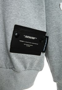 Sudadera gris con tejido texturizado, que presenta un parche de cuero negro con texto en relieve y dos botones a presión en el lateral.