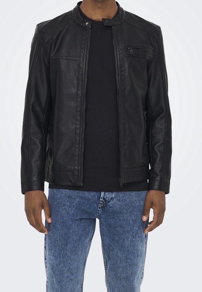 Giacca nera in pelle con colletto a listino, zip frontale e piccola tasca sul petto, indossata sopra una camicia nera e jeans di denim blu.