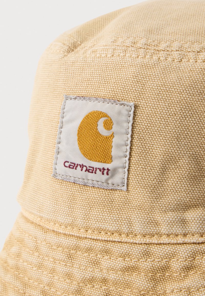Cappello a secchiello beige Carhartt in primo piano che mostra la trama del tessuto cucito e un'etichetta rettangolare con logo giallo e nome del marchio.