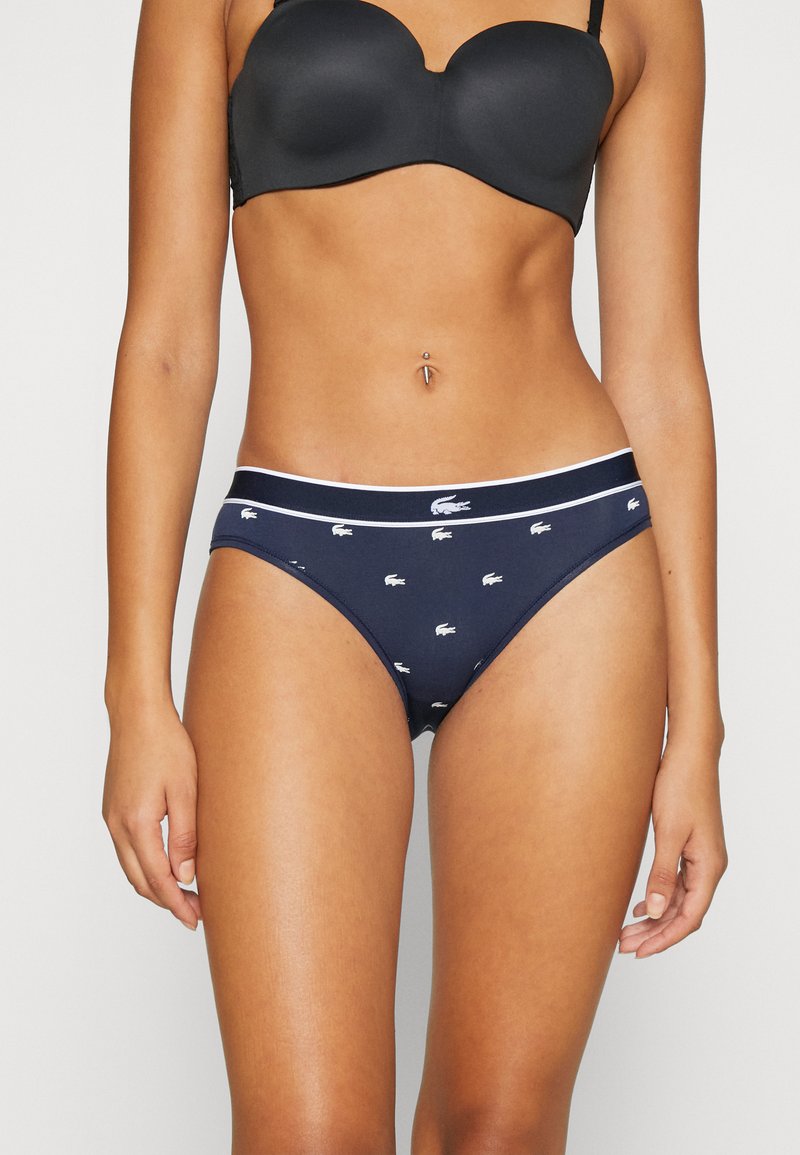 Jūras spēku kokvilnas bikini apakšbikses ar baltu Lacoste loga rakstu un elastīgu jostasvietu. Gluda tekstūra un klasiskā griezuma dizains.
