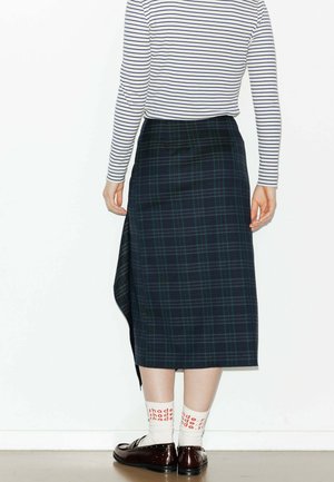 Jupe midi à carreaux sombres avec des motifs verts et bleus, fente sur le côté, portée avec des chaussettes blanches comportant du texte rouge et des chaussures marron. Haut à rayures verticales.
