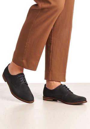 Chaussures oxford en suède noir à bouts ronds, avec trois lacets et une semelle marron. Portées avec un pantalon droit marron sur un fond neutre.