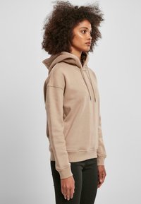 Beige hettegenser med snøring i hetten, ribbestrikkede mansjetter og linning, laget av myk stoff. Brukt med svarte tettsittende bukser, med sømløst design.
