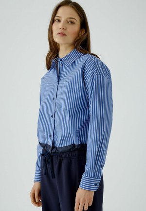 Jonge vrouw draagt een blauw-wit verticaal gestreept overhemd met knopen en donkere broek met trekkoord tegen een effen achtergrond.