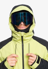 Gul och svart vattentät jacka med hög krage, dragkedjeslutning och justerbara dragsnören, bärs med stora spegelglasögon för skidåkning.