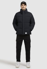 Must švaibiga jope kõrge kaelusega, tikandiga disain ja kaks külgtaskut. Kombineeritud mustade kargopükstega ja valgete tossudega.