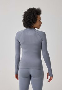 Lange mouwen actieve kleding top in lichtgrijs, met een aansluitend ontwerp, gestructureerde stof en duimopeningen bij de manchetten. Subtiele branding op de rug.