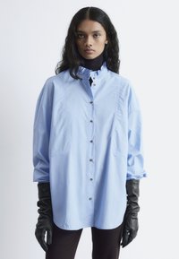 Light blue oversized button-up shirt with ruffled collar, featuring two side pockets and black leather gloves. Smooth fabric texture.