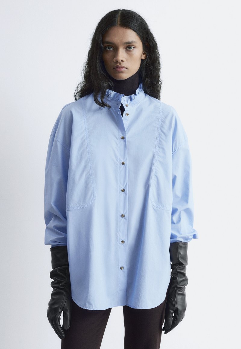 Light blue oversized button-up shirt with ruffled collar, featuring two side pockets and black leather gloves. Smooth fabric texture.