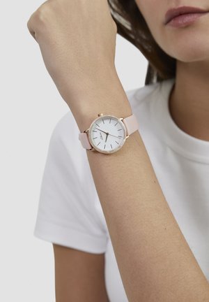 Ronde horloge met een witte wijzerplaat, gouden accenten en een roze siliconen band, gedragen om een pols tegen een effen achtergrond.
