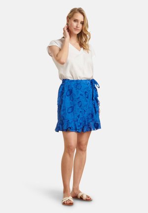 Blauwe rok met bloemmotief, voorzien van een geschulpte zoom en een strik aan de zijkant, gecombineerd met een witte blouse met v-hals en platte sandalen. Gladde stoftextuur.