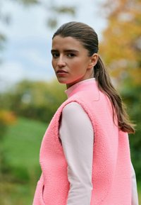 Roza flis telovnik z bočnimi žepi, oblečen preko svetlo rožnate majice z dolgimi rokavi, visoki ovratnik in teksturirano tkanino.