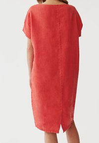 Frau in einem lockeren, knielangen, kurzärmeligen korallenfarbenen Kleid, von hinten gezeigt, mit einer Mittelnaht und einem kleinen Schlitz hinten.