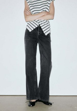 Personne portant un haut sans manches rayé, un jean noir à jambes larges et des ballerines noires, debout, les bras croisés devant un fond uni.