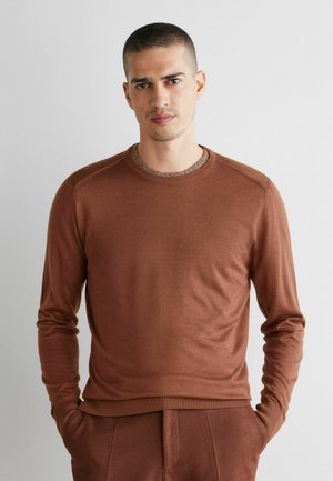 Mann mit kurzen Haaren, der einen braunen Pullover mit langen Ärmeln und passende braune Hosen trägt, steht mit den Händen in den Taschen vor einem einfarbigen Hintergrund.