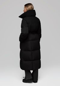 Žena s blond vlasy, oblečená v dlouhém černém péřovém kabátu s vysokým límcem, černých kalhotách a černých botách, stojící na jednobarevném pozadí.