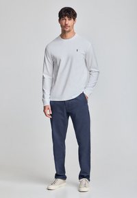 Witte katoenen t-shirt met lange mouwen en een klein zwart logo op de borst, gecombineerd met marineblauwe broek en lichtgekleurde sneakers.