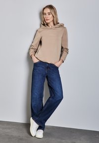 Sudadera con capucha beige hecha de material suave, combinada con jeans azul oscuro y zapatillas blancas. El modelo está de pie en un entorno neutro.