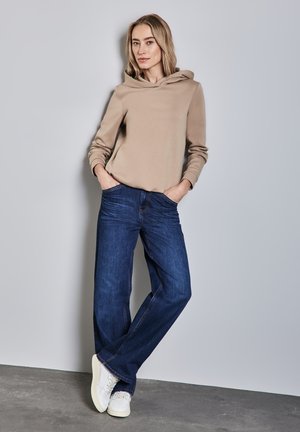 Sweatshirt à capuche beige en matière douce, associé à un jean bleu marine et des baskets blanches. Le modèle se tient dans un cadre neutre.