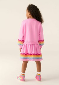 Rosa Kleid mit langen Ärmeln, das bunte Zickzackmuster an den Manschetten und dem Saum aufweist. Kombiniert mit rosa, knöchelhohen Sneakers.