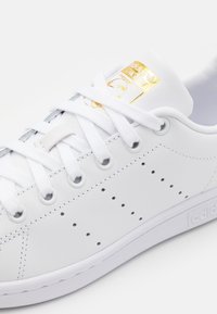 Vita läder sneaker med platt sula, snörningsdesign, perforerade detaljer och en guldfärgad logotyp på tungan. Mjuk textur.
