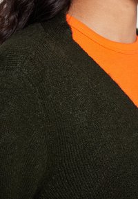 Grüner Strickpullover mit strukturierter Oberfläche, kombiniert mit einem leuchtend orangefarbenen Rundhalsausschnitt, der am Kragen sichtbar ist.