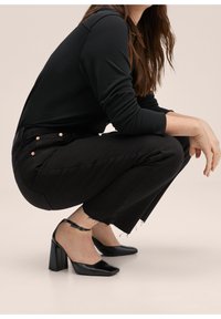 Top negro de manga larga, jeans negros de talle alto con dobladillo deshilachado y tacones negros de punta cuadrada con correa en el tobillo. Diseño simple y ajustado.