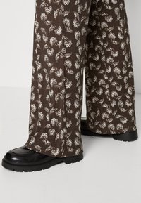 Hnědé kalhoty s vzorem a bílými květinovými motivy, vyrobené z texturované tkaniny. Vybavené lesklými černými mokasínami s masivní podrážkou.