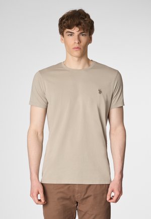 Un modello maschile si trova di fronte a uno sfondo semplice, indossando una maglietta beige con un piccolo logo e pantaloni marroni. La sua espressione è neutra.