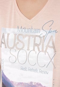 Różowa bawełniana koszulka z dekoltem w serek, z graficznym nadrukiem z napisem "Austria SOCSX" ozdobionym cyrkoniami oraz tekstem na tle górskiego krajobrazu.