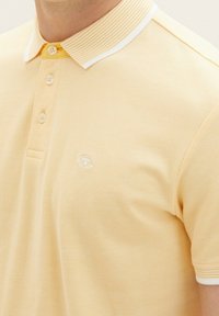 Polo en jaune clair, en tissu texturé. Caractéristiques d'un col rayé avec un bord blanc et trois boutons, logo brodé sur la poitrine.