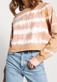 Oříznutá hnědá batikovaná mikina s bílými vzory, s dlouhými rukávy a měkkou texturou, stylizovaná s modrými vysoko sedícími džínami.