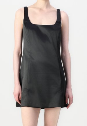 Kobieta w czarnej, satynowej, mini sukience bez rękawów z kwadratowym dekoltem na jednolitym białym tle.