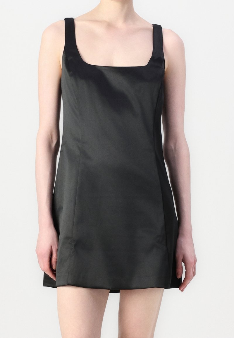 Femme portant une mini-robe noire en satin sans manches avec un col carré sur un fond blanc uni.