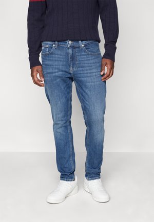 Jean en denim bleu avec une coupe slim, un design à cinq poches et un léger délavage. Accompagné de baskets blanches et d'un pull en tricot côtelé noir.