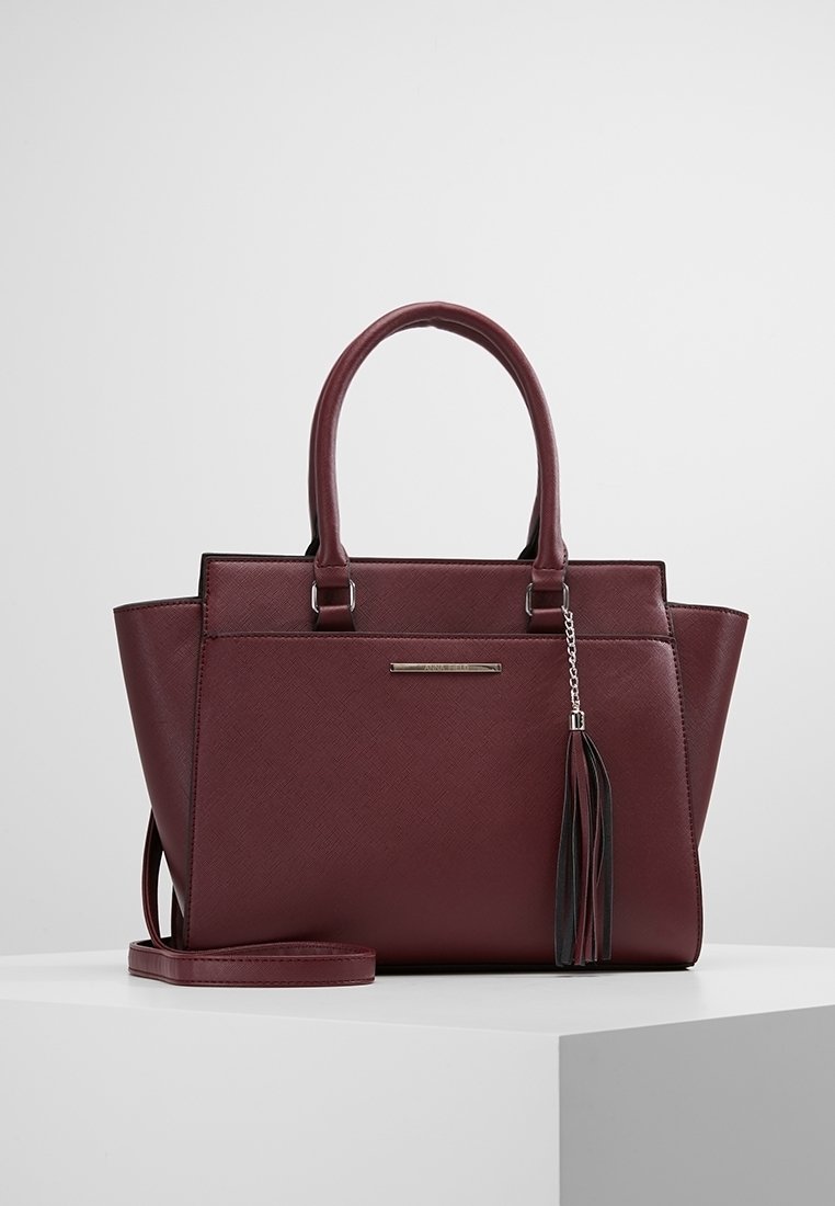 Sac à main en cuir rouge foncé avec doubles poignées, un pompon décoratif à l'avant et une bandoulière amovible sur une surface blanche.