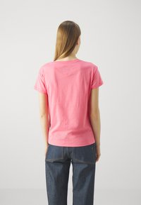 Różowa bawełniana koszulka z krótkim rękawem, okragłym dekoltem i kwadratową łatą na górnej części pleców. Uzupełniona niebieskimi dżinsowymi spodniami.