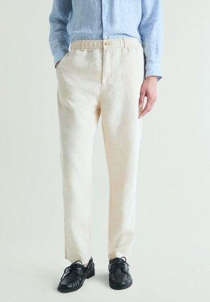 Persona que lleva pantalones beige de pierna recta, una camisa azul claro abotonada y zapatos negros con cordones, de pie con una mano en el bolsillo.