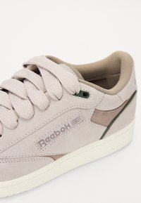 Light grey suede sneaker with cream rubber sole, featuring textured laces, green eyelet, and a subtle beige accent on the sides.