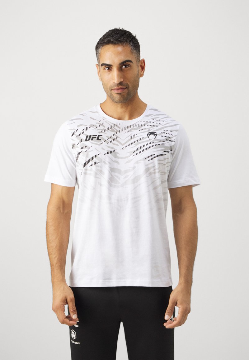 Camiseta blanca de manga corta con gráficos abstractos en negro en la parte delantera. Presenta el logo de UFC a la izquierda, un escote redondo y un corte holgado.