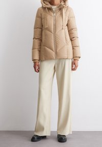 Beige Daunenjacke mit Kapuze, Reißverschluss vorne und gerippten Bündchen. Kombiniert mit weit geschnittenen cremefarbenen Hosen und schwarzen klobigen Stiefeln. Texturierte Oberfläche.