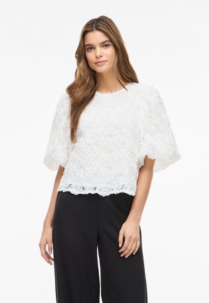 Ung kvinne med langt brunt hår iført en hvit, teksturert bluse med blomster og korte ermer, og svarte bukser med vide ben, stående foran en hvit bakgrunn.