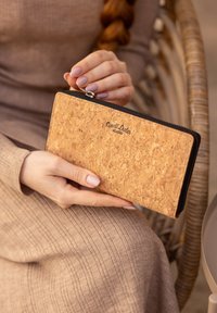 Kurkportemonnee met een gestructureerd oppervlak, ritsluiting en zwarte afwerking. Bedrukt logo aan de voorkant, vastgehouden tegen een geribbeld stoffen achtergrond.