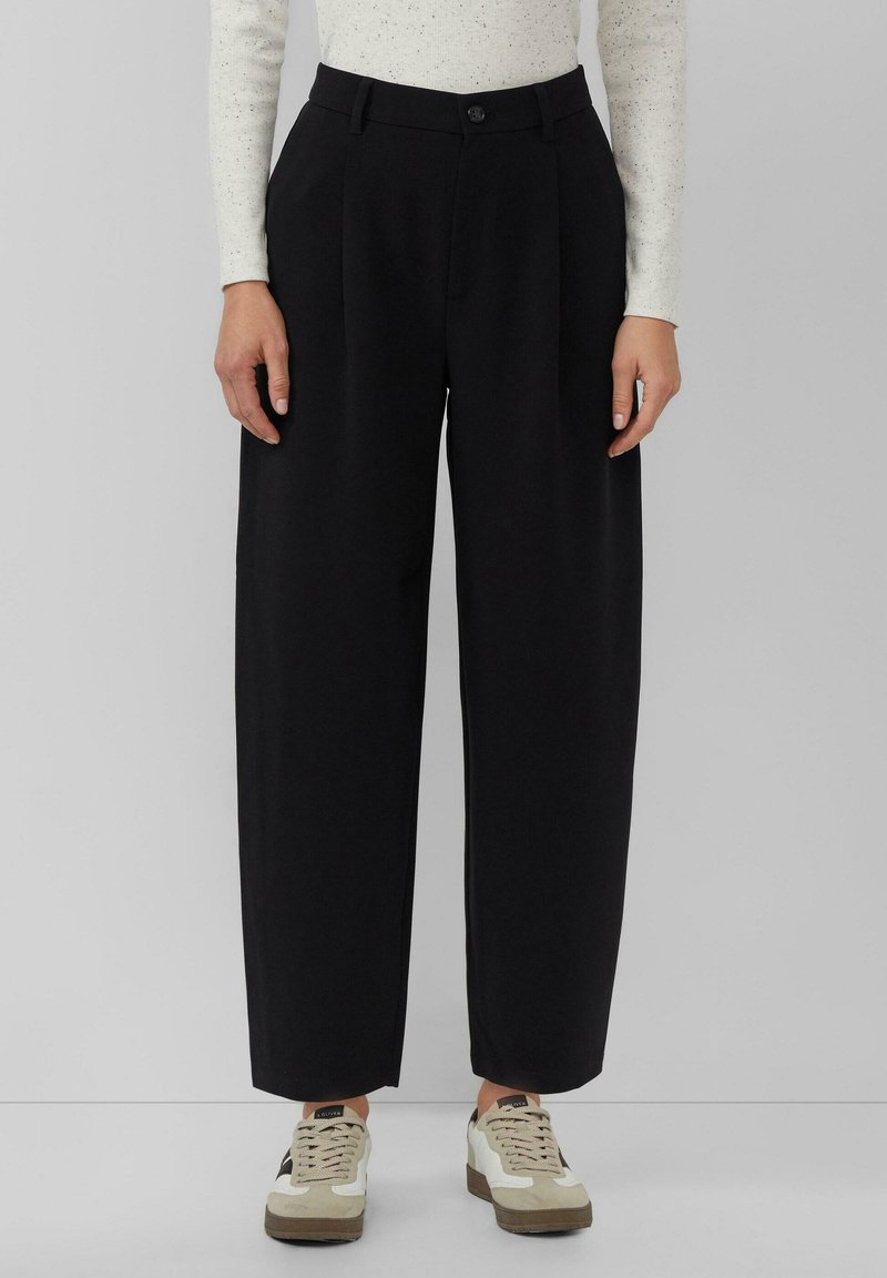 Zwarte broek met wijde pijpen en een hoge taille, voorzien van plooien aan de voorkant en een knoopsluiting, gecombineerd met beige-witte sneakers.