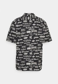 Chemise noire à manches courtes avec logos blancs répétés de la marque Dickies et phrases telles que "100 years of hard work" et "Quality Workwear."