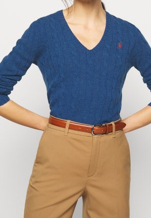 Femme portant un pull bleu à col en V et tricot torsadé, un pantalon taille haute couleur beige et une ceinture en cuir marron, les mains sur les hanches.