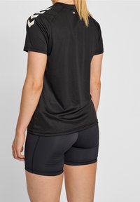 Musta urheilu t-paita lyhyillä hihoilla, jossa on valkoiset chevron-korostukset olkapäissä. Yhdistetty tummiin istuviin shortseihin. Sileä kankaan rakenne.