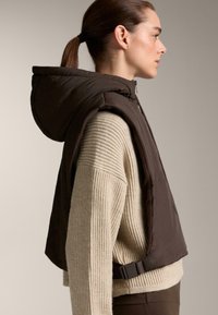 Hnědá puffer vesta s kapucí, zkrácený střih, s postranním přezkou a navrstvená přes béžový žebrovaný svetr. Hladká textura a detaily šití.