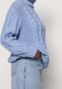 Maglione lavorato a maglia di colore azzurro chiaro con colletto alto, caratterizzato da motivi a treccia testurizzati e polsini a costine. Indossato sopra jeans in denim azzurro chiaro.